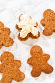 Gingerbread cookies await their second halves on a marble surface, each meticulously piped with buttercream to craft delightful sandwich treats. The precision of the piping adds a festive touch.