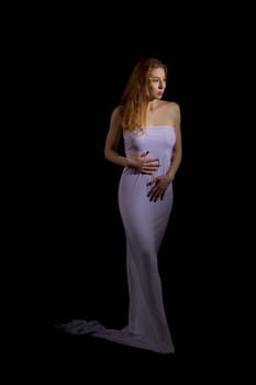 Delicate female with long hair and in white maxi dress standing on black background in studio and looking away