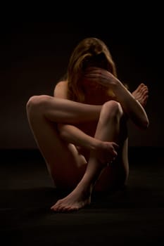 Sexy nude female with perfect body sitting difficult yoga pose in dark studio and hide face on knees