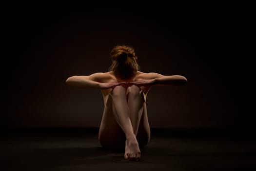 Sexy nude female with perfect body sitting on floor in dark studio and hide face on knees