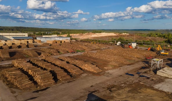 Drone view of scenery of piles of wooden logs stacked on ground in industrial area with contemporary vehicles at sawmill on sunny day