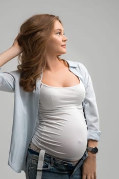 Delighted pregnant female in casual clothes standing on gray background and look a side