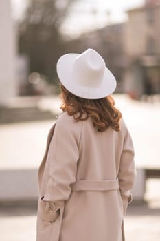 Back view of happy woman wearing hat and coat walking down street on sunny spring day. People, lifestyle, travel and vacations concept.