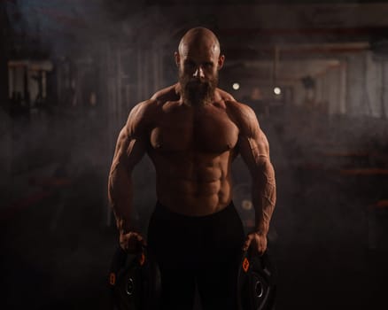 Muscular bald shirtless man holding weight plates. A bodybuilder shows off his form in the gym