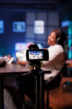 Male and female African American influencers are recording a podcast in neon-lit studio. They are using digital camera to create content for internet video channel and live streaming on social media.