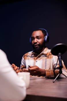 Black male host interviewing a person at home, using modern microphone and wireless headphones for his online talk show. African american man talking with an individual recording a podcast episode.