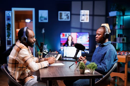 Sitting at table using the audio equipment are two african american men discussing and addressing their online audience. Interviewer asking male influencer questions, recording a radio show at home.