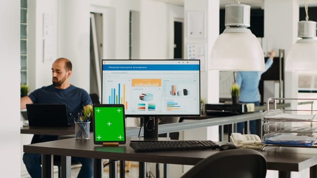 Empty desk with greenscreen on tablet, modern coworking space in creative agency office with employees at job. Blank mockup isolated template at workstation, small business agency.