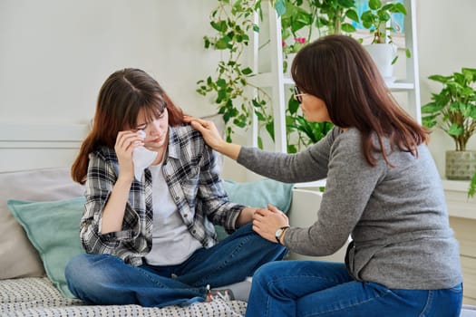 Upset sad crying daughter teenager sitting together with mother on couch at home. Family, communication, motherhood, friendship, relationship between parent and teen daughter concept