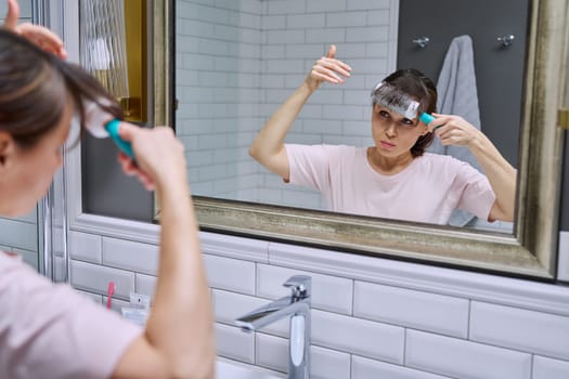 Middle-aged woman with comb looking in mirror, doing hair styling, in bathroom. Beauty, age, cosmetics, hair accessories