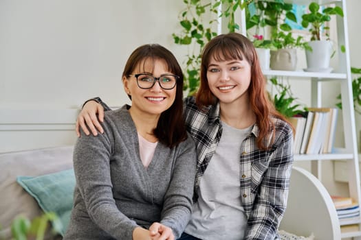 Portrait of happy hugging mother and teenage daughter sitting together on couch, smiling looking at camera. Two generations of family, communication, motherhood, friendship, relationship, mothers Day