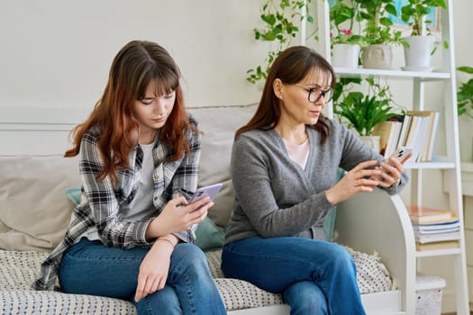 Mother and teenage daughter sitting on the couch at home, using smartphones. Technology, lifestyle, two generations of family, motherhood, relationship concept