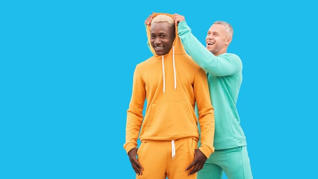 Multi ethnic friends. Two men different color black African-American ethnicity and white Caucasian ethnicity standing isolated blue background. One man put on hood to other male
