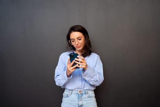 Pensive woman in glasses use smartphone, types text message on mobile phone, enjoys online communication, types feedback, wears denim shirt, isolated on black studio background. Technology concept