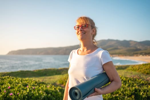 Elderly Caucasian lady wearing glasses holding yoga mat smiling on beach. Getting ready for morning yoga session on ocean shore, promoting healthy lifestyle and idea of enjoying retirement on vacation