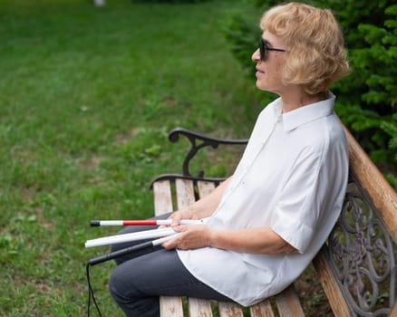 An elderly blind woman sits on a bench in the park with a folded tactile cane in her hands