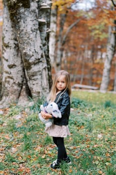 Little girl with a toy rabbit in her hands stands half-turned in the autumn forest. High quality photo