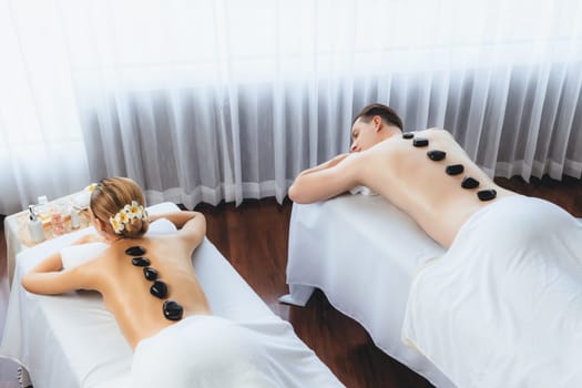 Hot stone massage at spa salon in luxury resort with day light serenity ambient, blissful couple customer enjoying spa basalt stone massage glide over body with soothing warmth. Quiescent