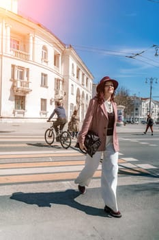 Woman city road crossing. Stylish woman in a hat crosses the road at a pedestrian crossing in the city. Dressed in white trousers and a jacket with a bag in her hands