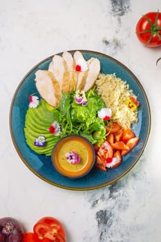Top view of nutritious lunch with grilled chicken, salad, tomatoes, avocado, couscous dipping sauce on blue plate.