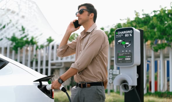 Young man put EV charger to recharge electric car's battery from charging station in city commercial parking lot. Rechargeable EV car for sustainable environmental friendly urban travel. Expedient