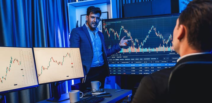 Two stock exchange traders discussing dynamic valued investment rate on monitor at night in panorama photo view. Businessman in stock market presenting economic in neon light at workplace. Sellable.