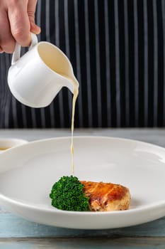 Chef pouring sauce over a dish prepared with grilled chicken breast and broccoli