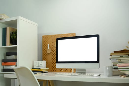 Workplace with blank computer screen and pile of book on white desk.