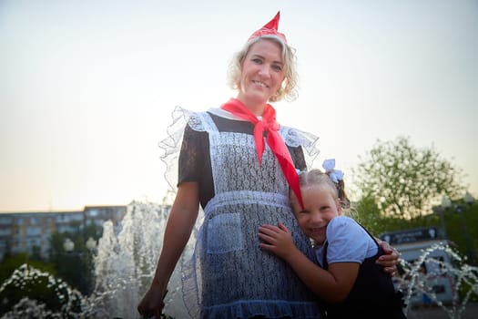 Young and adult schoolgirl on September with flowers having fun near water of fontain. Generations of schoolchildren, pioneer of USSR and October girl in modern uniform of Russia. Mom and daughter