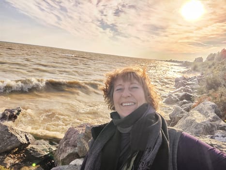 Happy cheerful middle aged woman in scarf taking selfie on nature outdoors and sun with water and waves of sea on the background