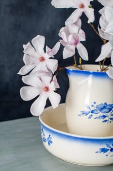 A branch of a delicate pink magnolia flower in full bloom in a vintage ceramic jug for washing on a dark background, spring still life, high quality photo