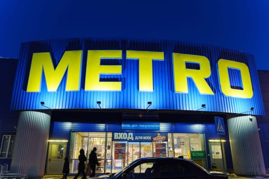 Tyumen, Russia-March 18, 2024: Metro sign glows brightly against the night sky, with the silhouettes of city buildings in the background.