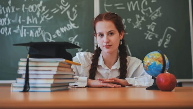 A red-haired high school senior poses against a backdrop of books, a globe and a graduation cap