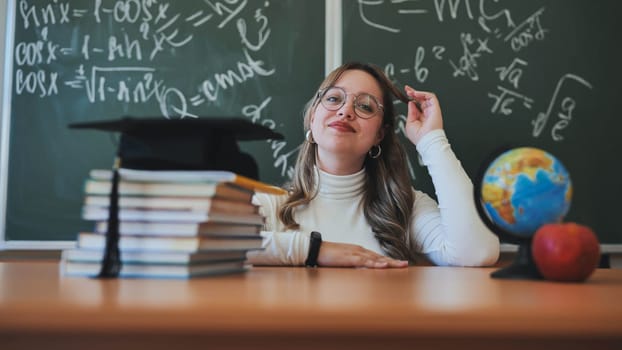 A schoolgirl wearing glasses poses against a background of books, an apple, a globe and a graduation cap