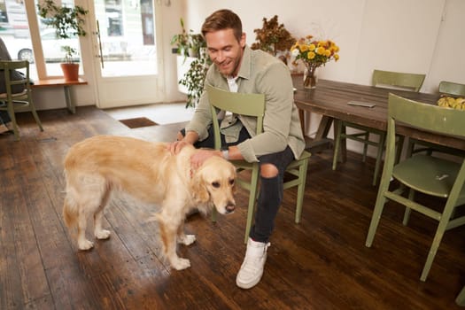 Close up portrait of handsome young man waiting for his order in coffee shop, petting a dog, resting in pet-friendly cafe.