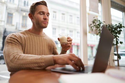 Portrait of handsome young man sitting in cafe with laptop. Digital nomad, freelancer working outdoors from coffee shop, typing something.