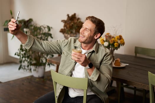 Portrait of handsome happy young man, taking selfie in cafe, posing with cup of coffee, video chats using smartphone app, talks to a friend. Lifestyle and leisure concept