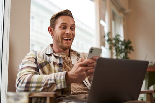 Image of man in cafe, coffee shop visitor sitting in chair with laptop and smartphone, looking surprised and excited at mobile phone screen, amazed by big news, promo offer on app.