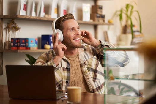 Lifestyle portrait of smiling digital nomad, man in cafe, working on laptop, listening music in wireless headphones and smiling, dancing on chair while enjoying favourite song.