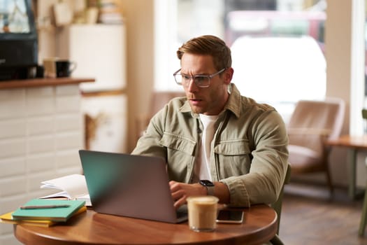 Portrait of man with serious face, sitting in cafe, wearing glasses, working on laptop, looking concentrated, doing freelance project.