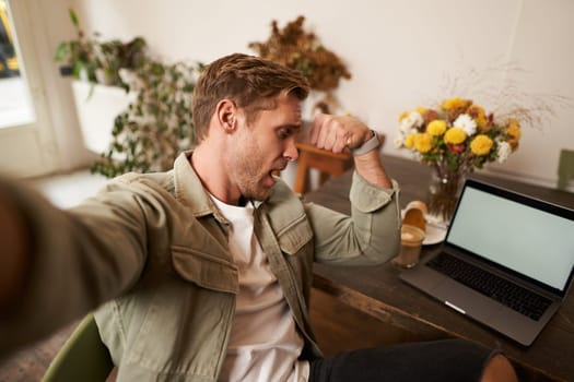 Man takes selfie on mobile phone, flexing biceps, shows his muscles at camera, makes photos in a cafe, sits in front of laptop.