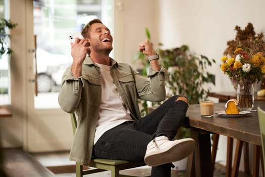 Portrait of relieved, happy young man, listens to happy news or podcast in earphones, sits in coffee shop, holds smartphone, raises hands up and smiling from excitement, spending time in cafe.