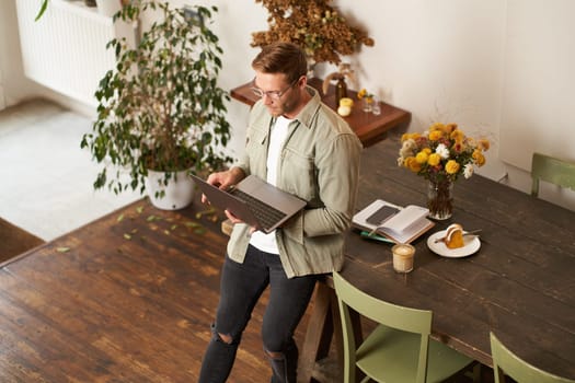 Image of young man, successful businessman sitting on table, looking at laptop, working in an office, looking concentrated on the project, doing job task, drinking coffee.