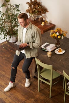 Portrait of handsome young man sits with laptop, looks at screen with concentrated, thoughtful face, working on a project, e-learning at co-working space, drinking coffee.