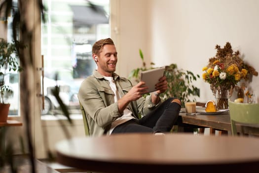 Portrait of handsome happy guy, young man sits in cafe, watching videos on digital tablet, wearing wireless headphones, laughing and smiling, spending time in coffee shop.