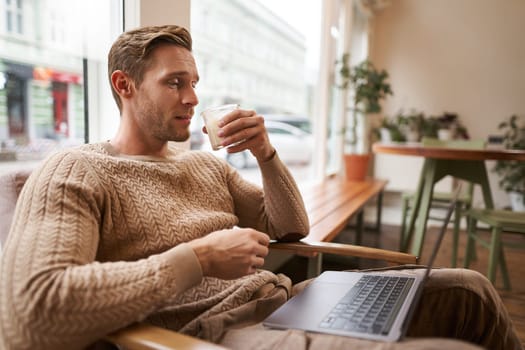 Working people and public space concept. Young handsome man, ux ui designer sitting with laptop in cafe, drinking cappuccino, looking at screen with serious face.