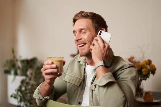 Portrait of young handsome man talking on the phone, drinks coffee and laughs. A guy sits in cafe having a nice, pleasant conversation over cup of cappuccino.