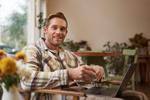 Handsome young professional, businessman sitting in cafe with glass of coffee and working on laptop, searching for inspiration outside of office workspace.