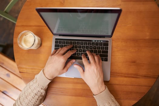 Close up portrait of male hands typing on keyboard, working on laptop in cafe, computer and cappuccino standing on wooden table in coworking space.