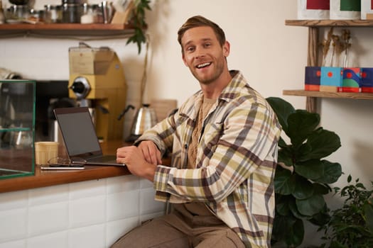 Small business concept. Handsome businessman, cafe owner, sitting near counter with laptop and smartphone, working in coffee shop.